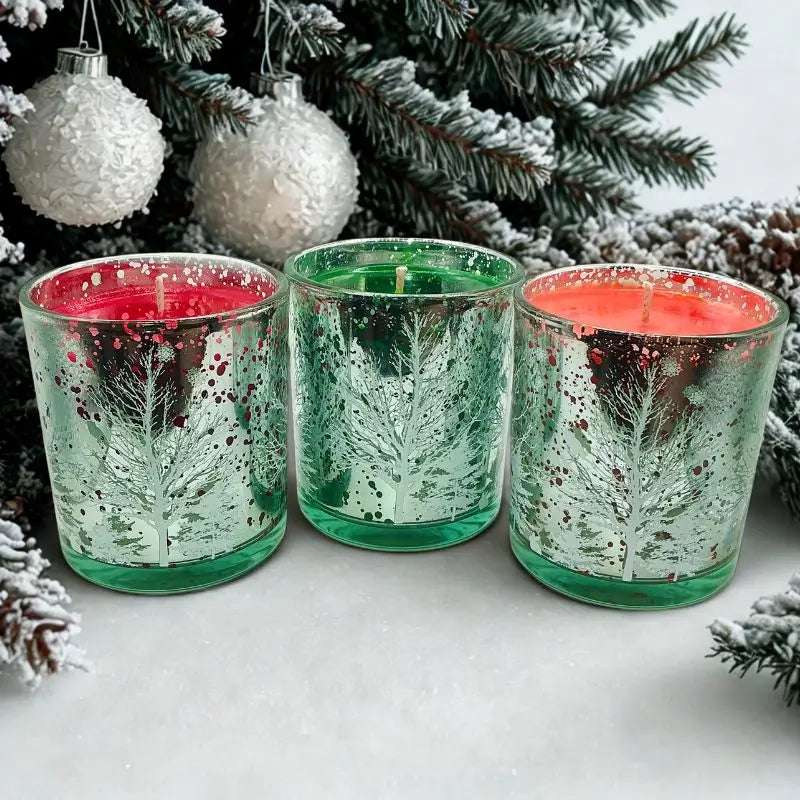 Three Christmas Candles in Decorative Glass with festive tree designs beside snow-covered branches.