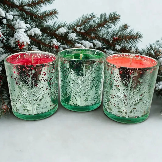 Christmas Candles in decorative glass holders with festive tree designs, placed in a snowy setting.