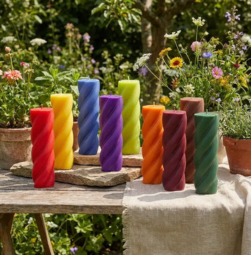 Customisable Candle in various colors arranged outdoors with flowers and greenery.